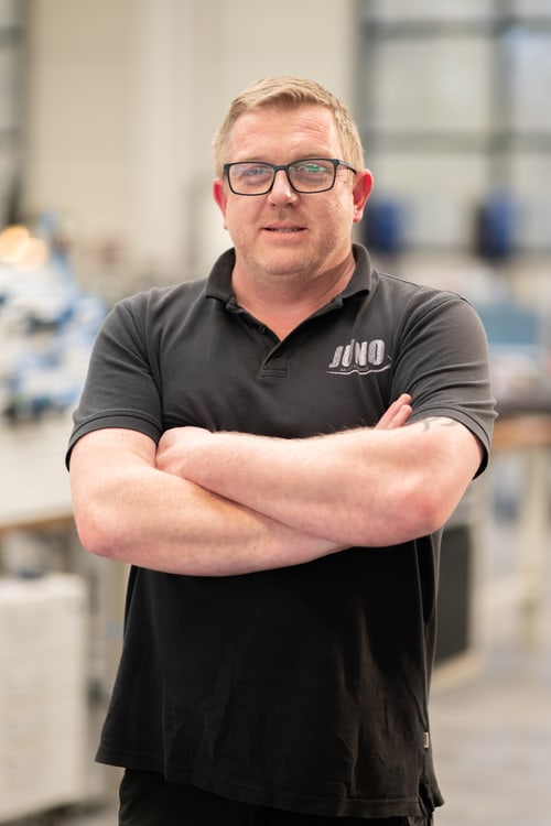 Mitarbeiter mit Brille im schwarzen JUNO-Poloshirt, Arme verschränkt, in der Werkhalle