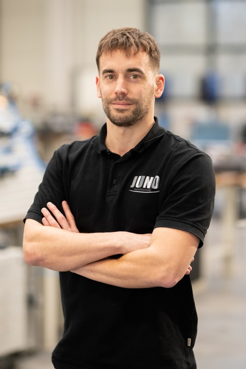 Mitarbeiter mit kurzem Haar im schwarzen JUNO-Poloshirt, Arme verschränkt, in der Werkhalle