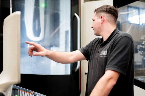 Mann in schwarzem Poloshirt bedient CNC-Maschine, zeigt auf den Bildschirm.